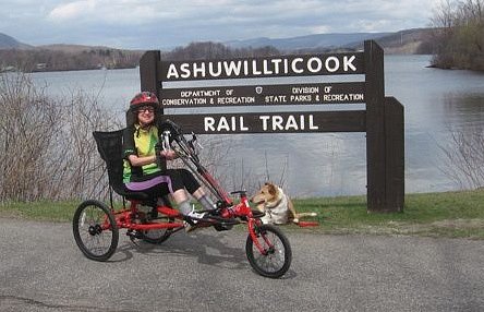Participant smiling and seated on an adapted trike in front of Ashuwillticook Rail Trail sign (with dog).