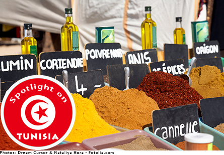 Spices in tubs at a spice market, bottles of olive oil are behind the spices. Red badge with Tunisian flag symbol of crescent moon with a star says, 