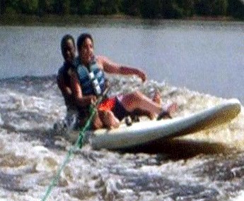 Two smiling people seated on a surfboard pulled like a waterski on the lake.