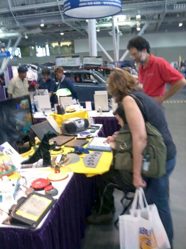 Tables of computer access technologies on display with people exploring. 