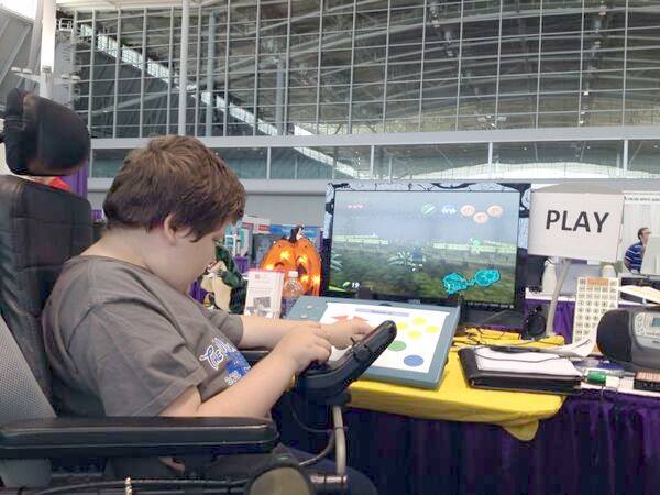 A young man in a power wheelchair using alternative computer access to play a game on a monitor. 