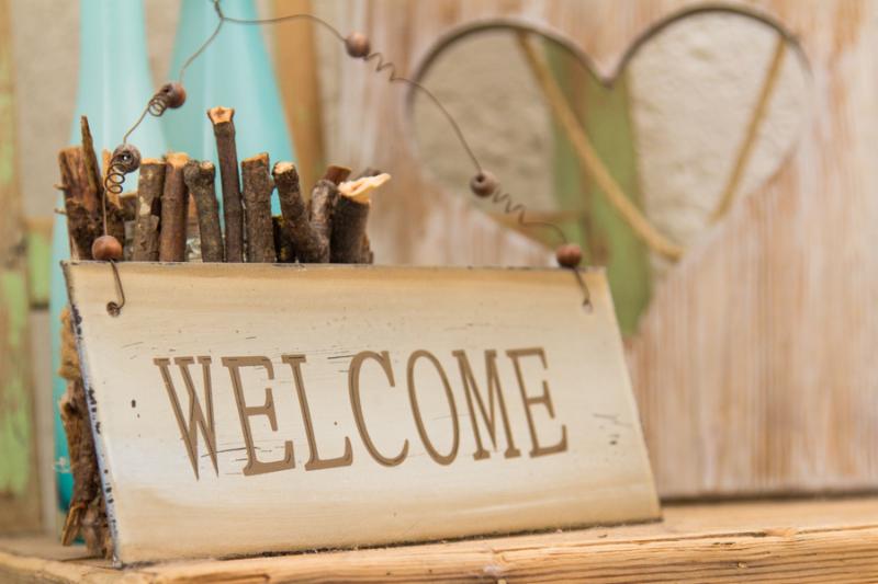 Rustic wooden WELCOME sign standing on a wooden shelf in front of a wood panel with a cut out heart