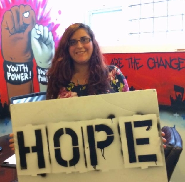 Brianna holding the Hope Board in front of the YP! mural of fists with the YOUTH POWER! wristband 