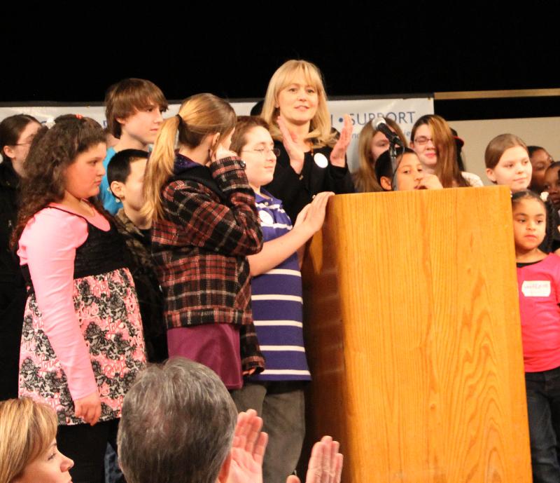 Kristin stands at the podium surrounded by children at the FTNYS legislative Lunch